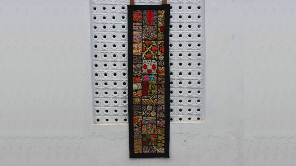 "Vertical boho wall hanging with colorful embroidered patchwork, framed in black, displayed against a white pegboard wall