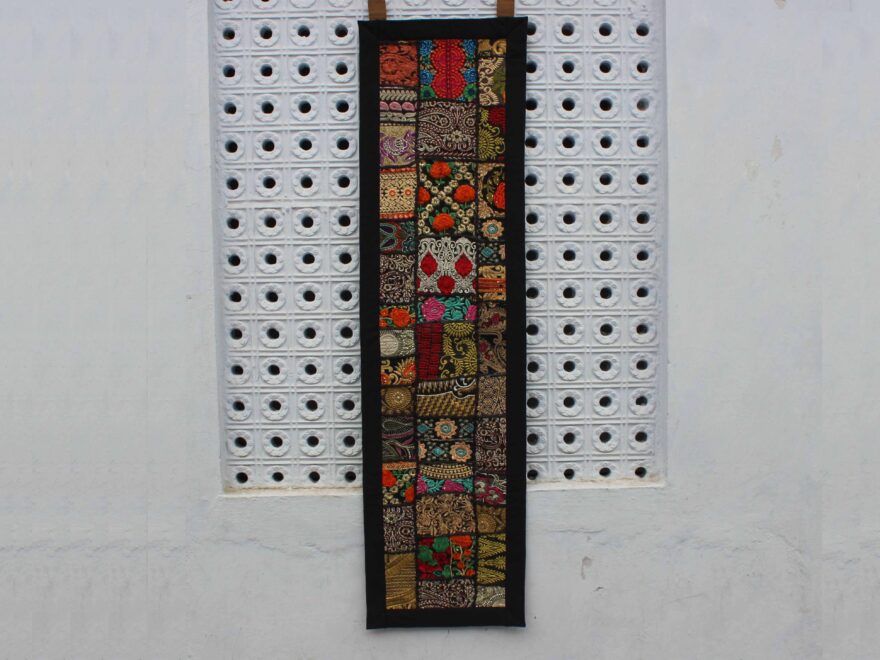 "Vertical boho wall hanging with colorful embroidered patchwork, framed in black, displayed against a white pegboard wall