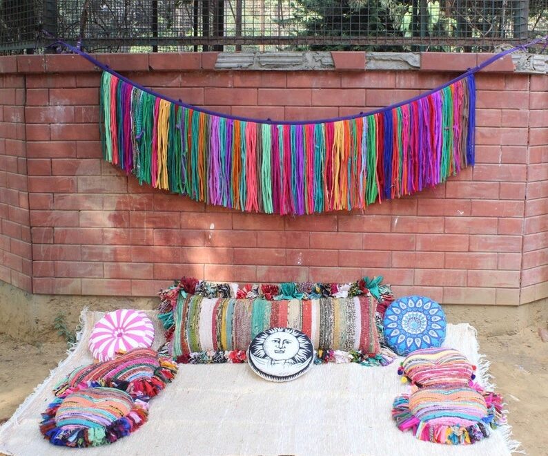 Colorful handmade boho valance made from yarn strands hanging on a brick wall, styled with eclectic embroidered pillows and rustic decor